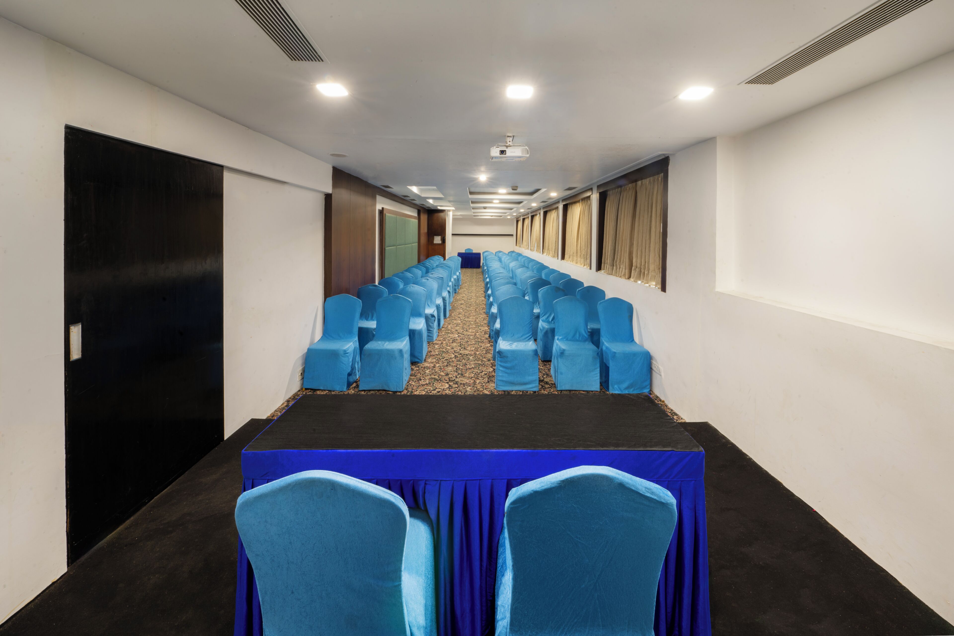 A conference room with rows of blue chairs, a black table, and a carpeted floor.