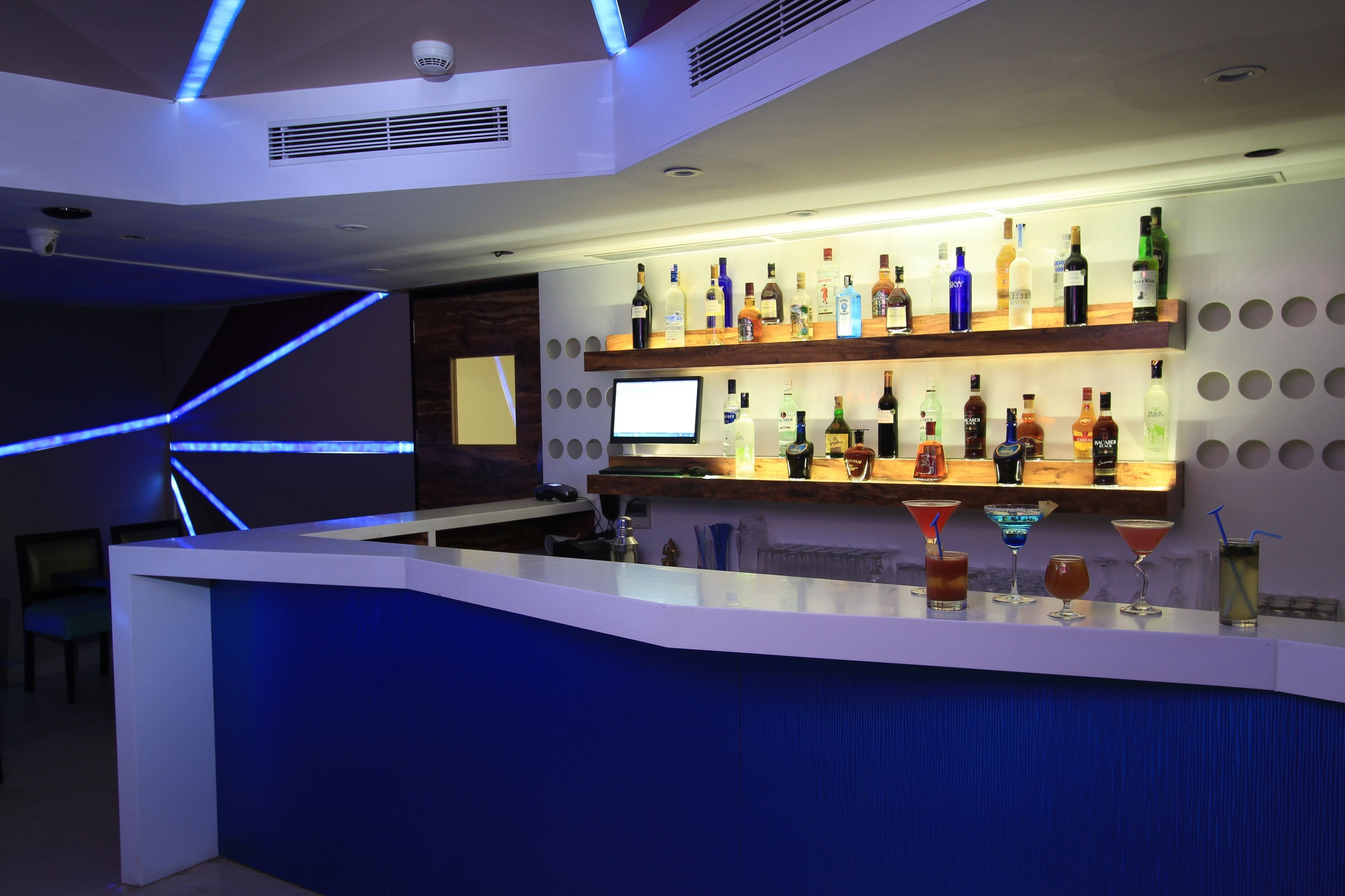 Uma área de bar moderna com balcão branco, prateleiras com diversas garrafas de bebidas e um monitor de computador.