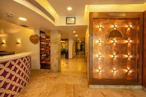 A hotel reception area with a marble floor, a reception desk with a patterned design, a 'Welcome' sign, and a wall with illuminated fan-shaped lights.