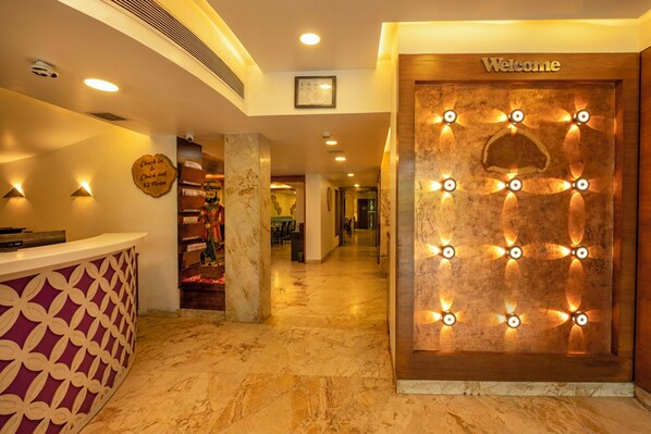 A hotel reception area with a marble floor, a reception desk with a patterned design, a 'Welcome' sign, and a wall with illuminated fan-shaped lights.