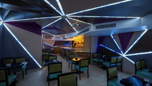 Un restaurante moderno con iluminación de techo geométrica, sillas tapizadas en verde y una zona de bar al fondo.