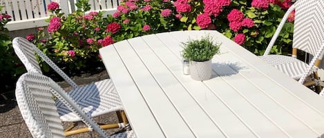 Ein weißer Gartentisch mit zwei Korbstühlen, umgeben von einem weißen Zaun und leuchtend rosa Blumen.