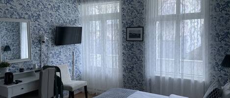 A bedroom with a bed, a desk, a chair, a TV, and a window with curtains.
