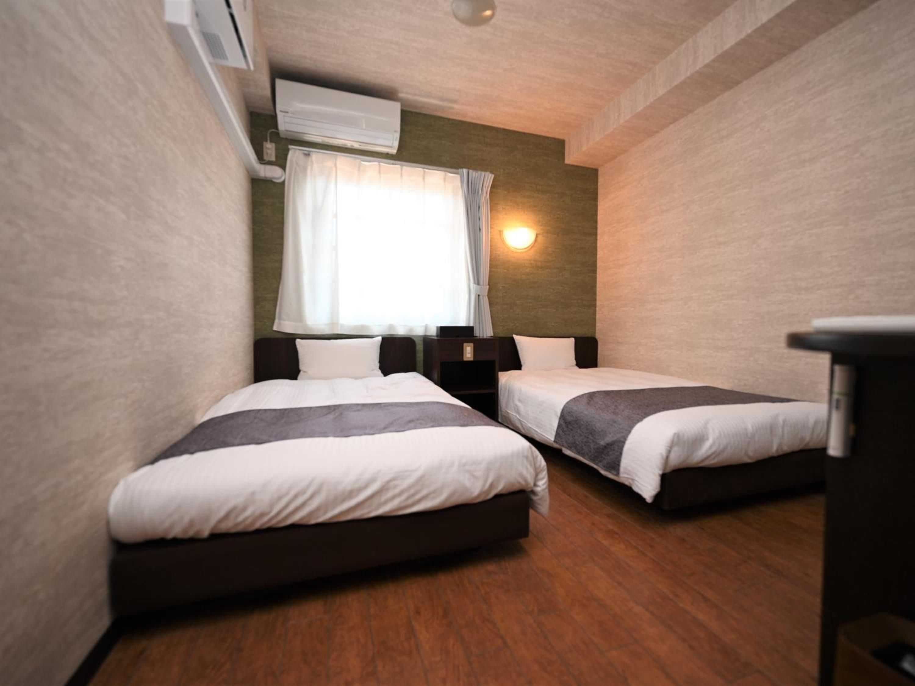 A hotel room with two beds, a window with curtains, a wall-mounted air conditioner, and a bedside table.