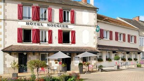 A hotel with a sign reading 'HOTEL NOUGIER,' featuring a traditional facade with red shutters, outdoor seating with umbrellas, and a paved courtyard. - Hôtel Nougier (Fursac)