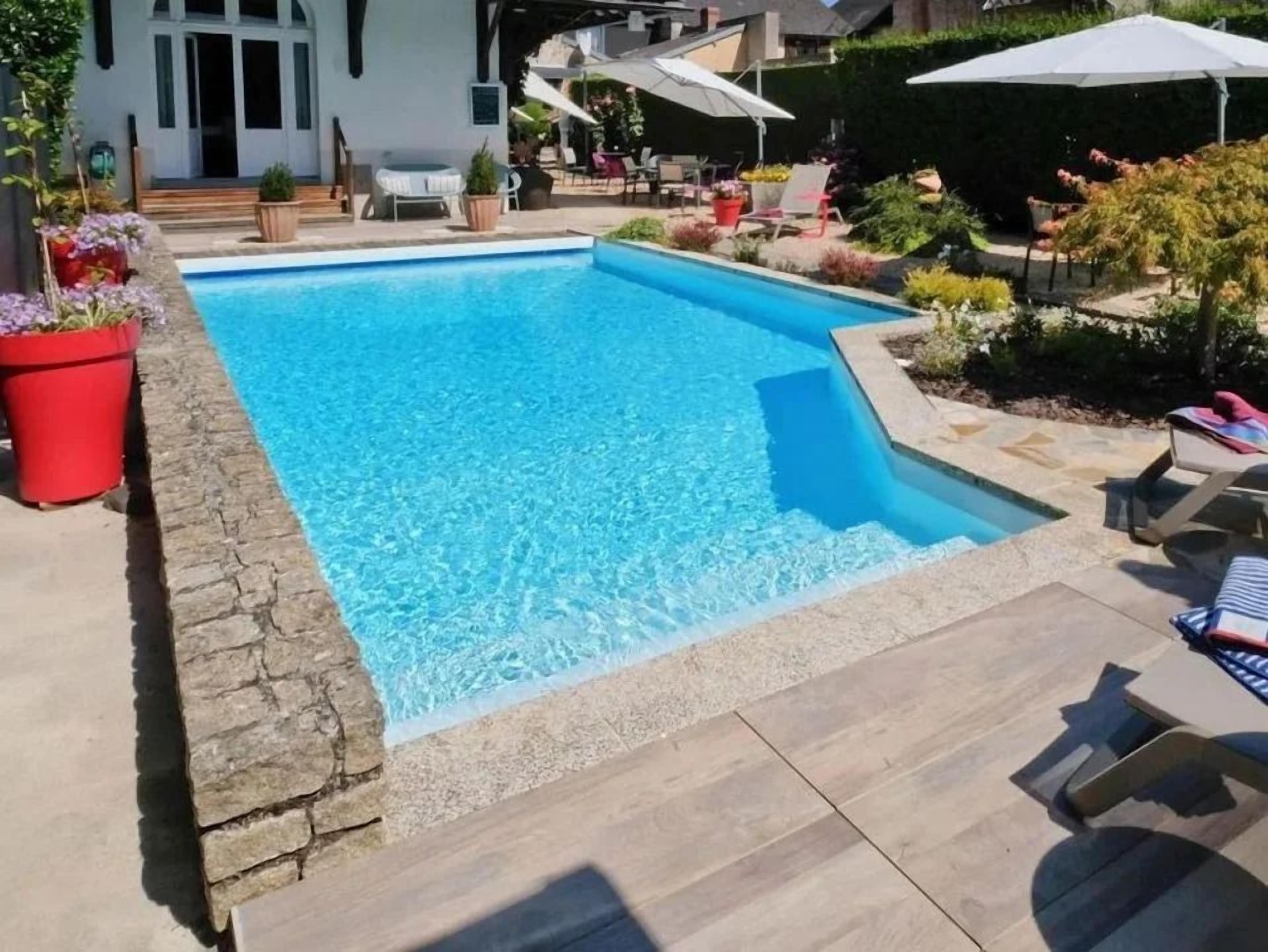 A rectangular swimming pool with a stone border, surrounded by a patio area with lounge chairs and umbrellas.