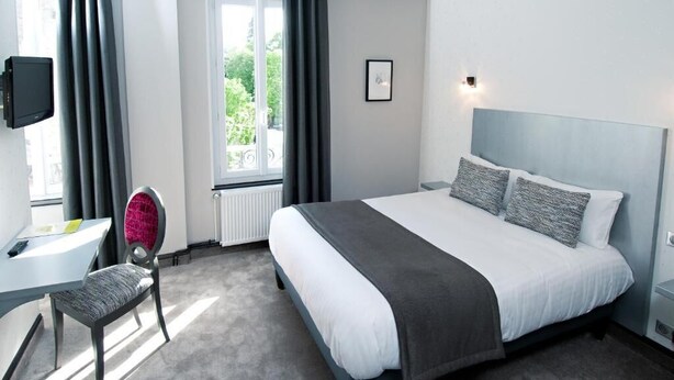 A hotel room with a bed, a desk, a chair, a TV, and a window with curtains. - Hôtel Nougier (Fursac)