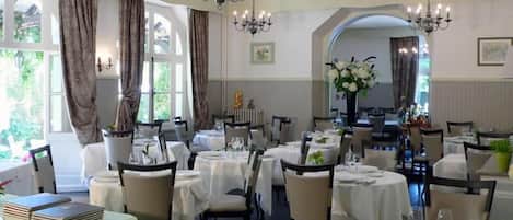 A dining area with tables set for service, chandeliers, and large windows with curtains.