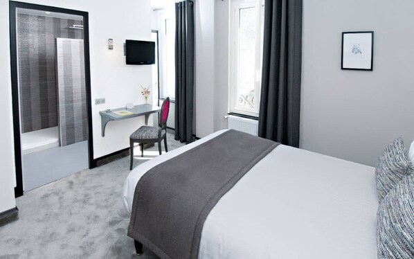 A hotel room with a bed, a desk, a chair, a TV, and a window with curtains. - Hôtel Nougier (Fursac)