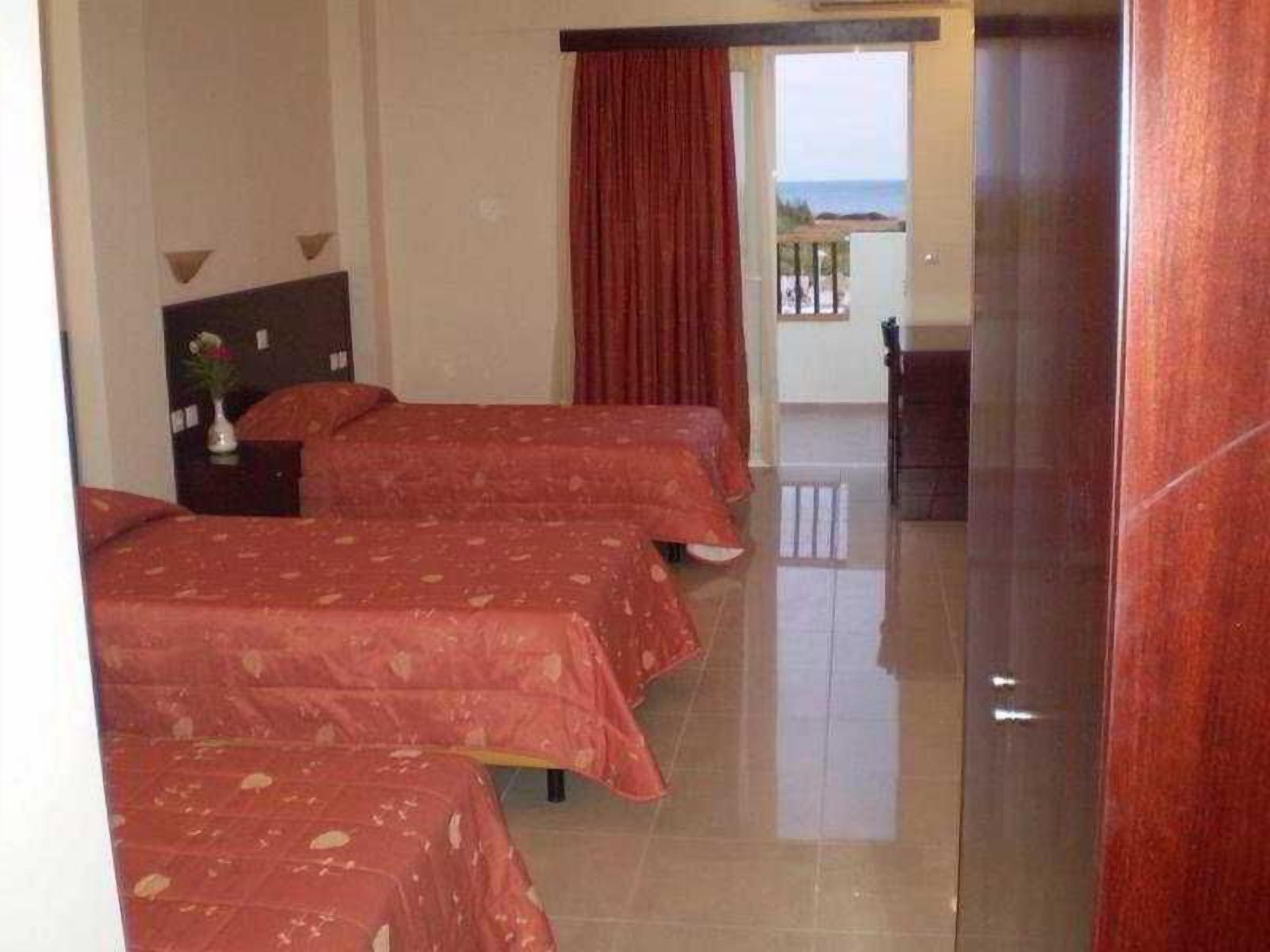 A hotel room with three beds, a wooden headboard, a nightstand with a vase, and a view of the sea through a balcony door.