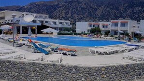 A swimming pool with lounge chairs and umbrellas, surrounded by a stone wall and buildings with a mountainous backdrop. - Evripides Village (Kos)