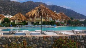 A pool area with lounge chairs and thatched umbrellas, surrounded by stone walls and mountains in the background. - Evripides Village (Kos)