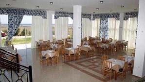 A spacious dining area with multiple tables and chairs, large windows with curtains, and a view of the landscape. - Evripides Village (Kos)