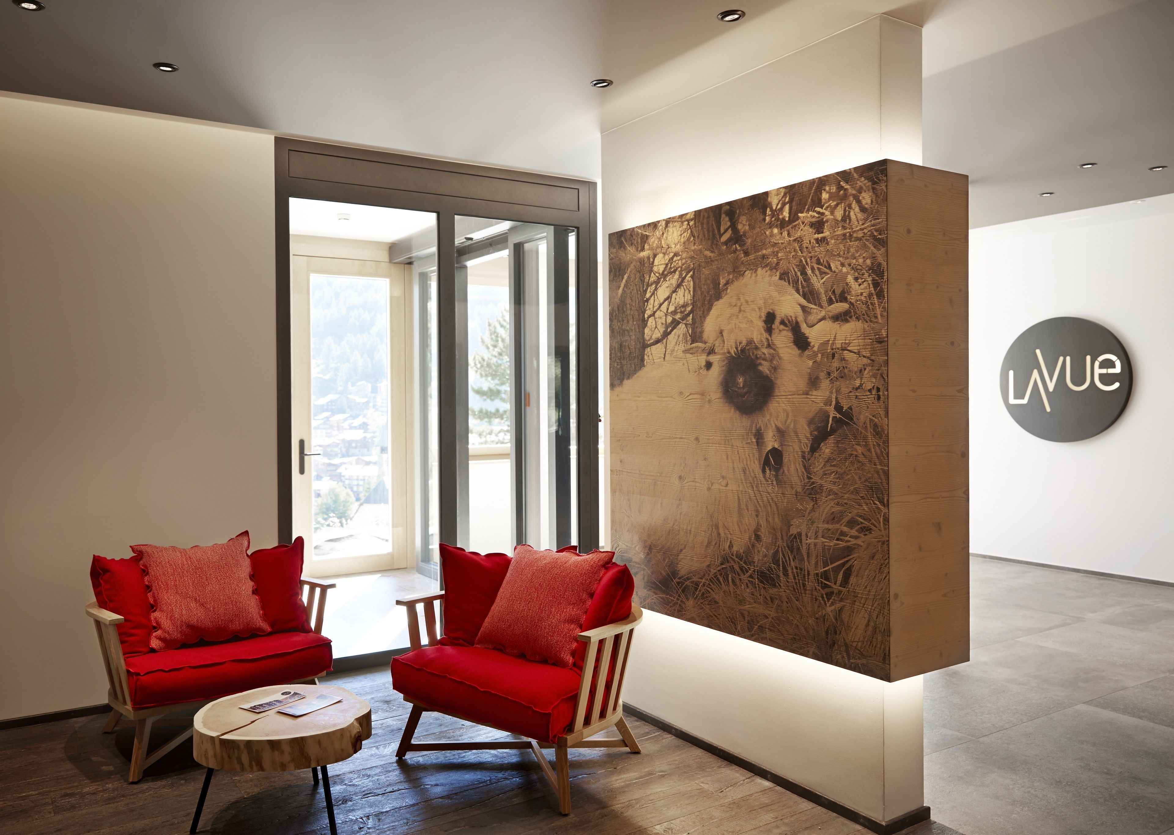 Ein modernes Interieur mit zwei roten Sesseln, einem runden Holztisch und einem großen, abstrakten Kunstwerk an der Wand.
