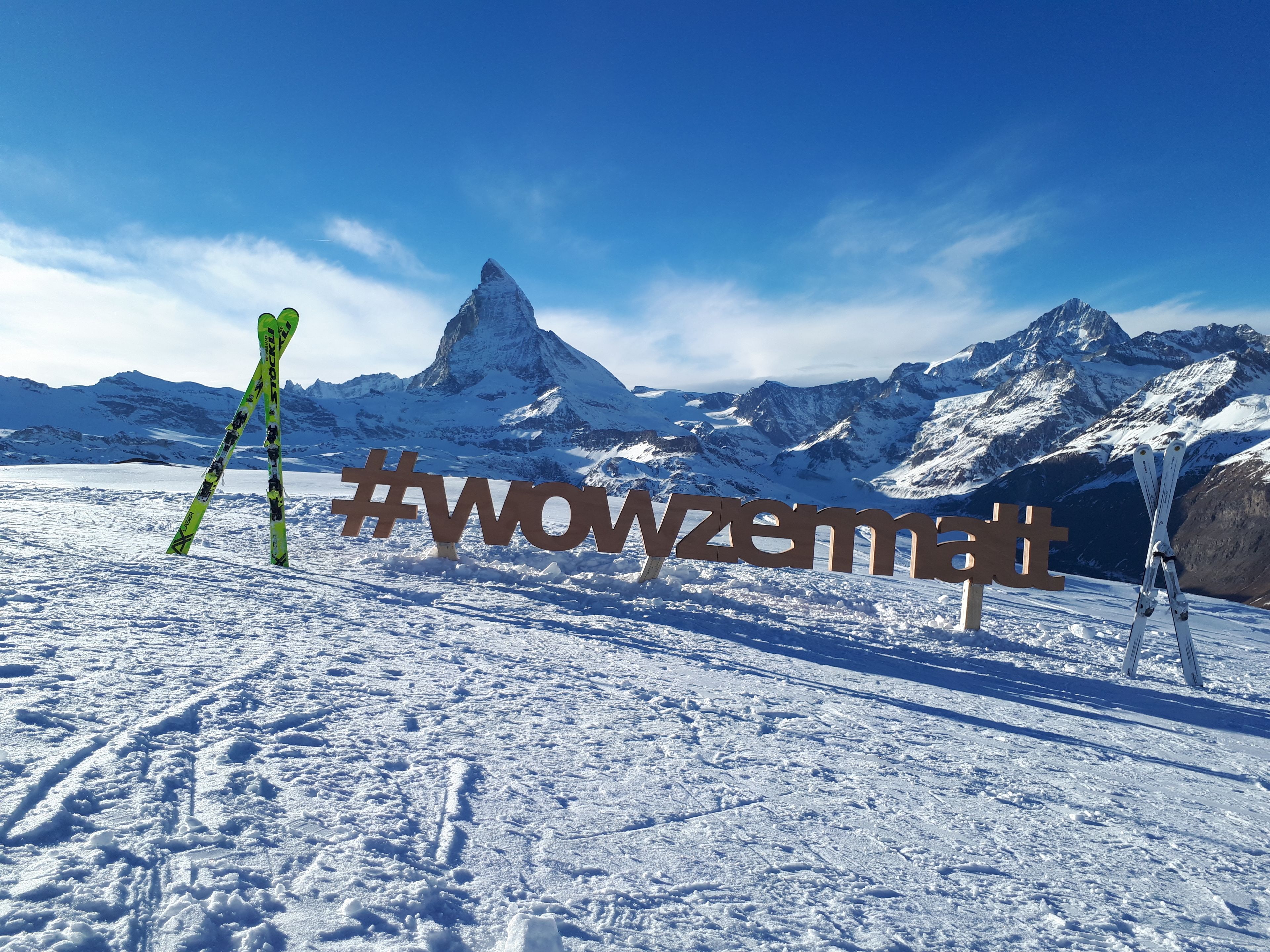 Eine verschneite Berglandschaft mit einem markanten Gipfel im Hintergrund, Skier, die an einem Holzschild mit dem Hashtag #wowzemt lehnen.