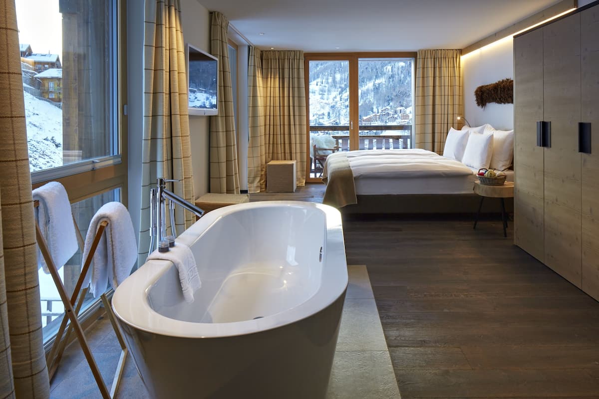 Habitación de hotel moderna con una bañera grande, una cama y un balcón con vistas a una montaña nevada.