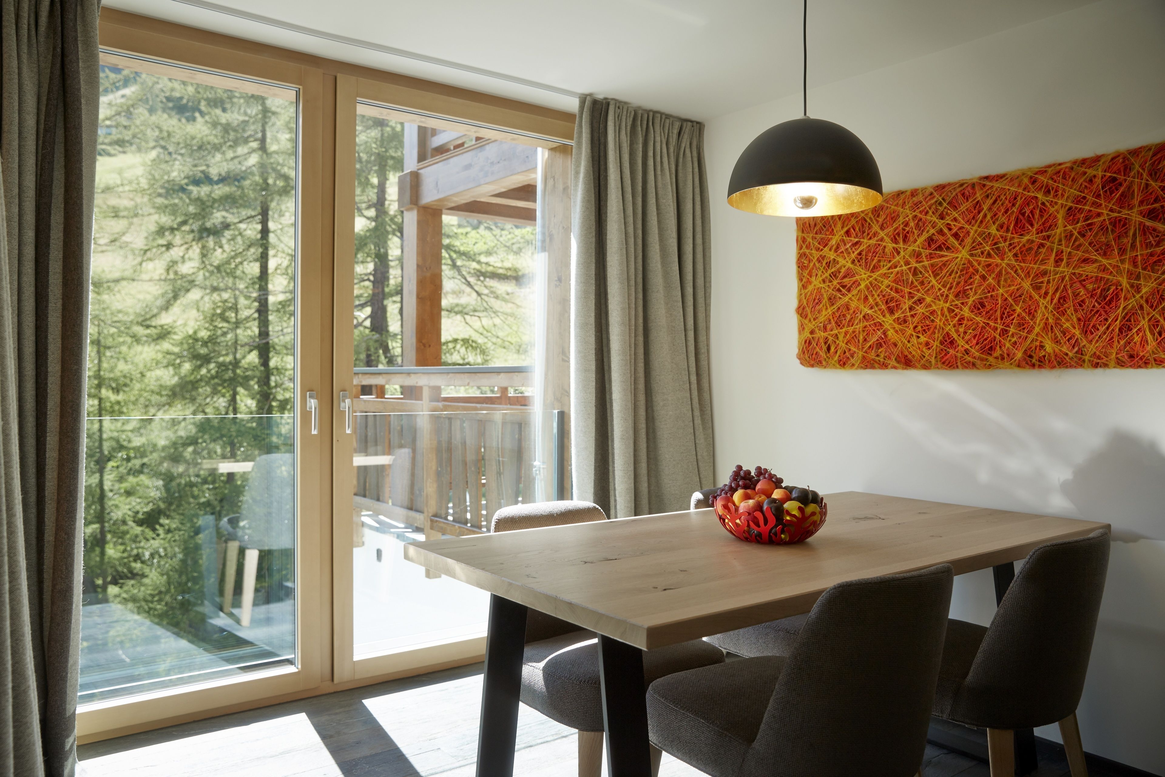 Ein Essbereich mit einem Holztisch, grau gepolsterten Stühlen, einer Obstschale, einer schwarzen Pendelleuchte und einem großen Fenster mit Vorhängen.