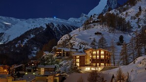 A snow-covered mountain with a prominent peak, a wooden building with multiple balconies, and several smaller structures in the foreground.