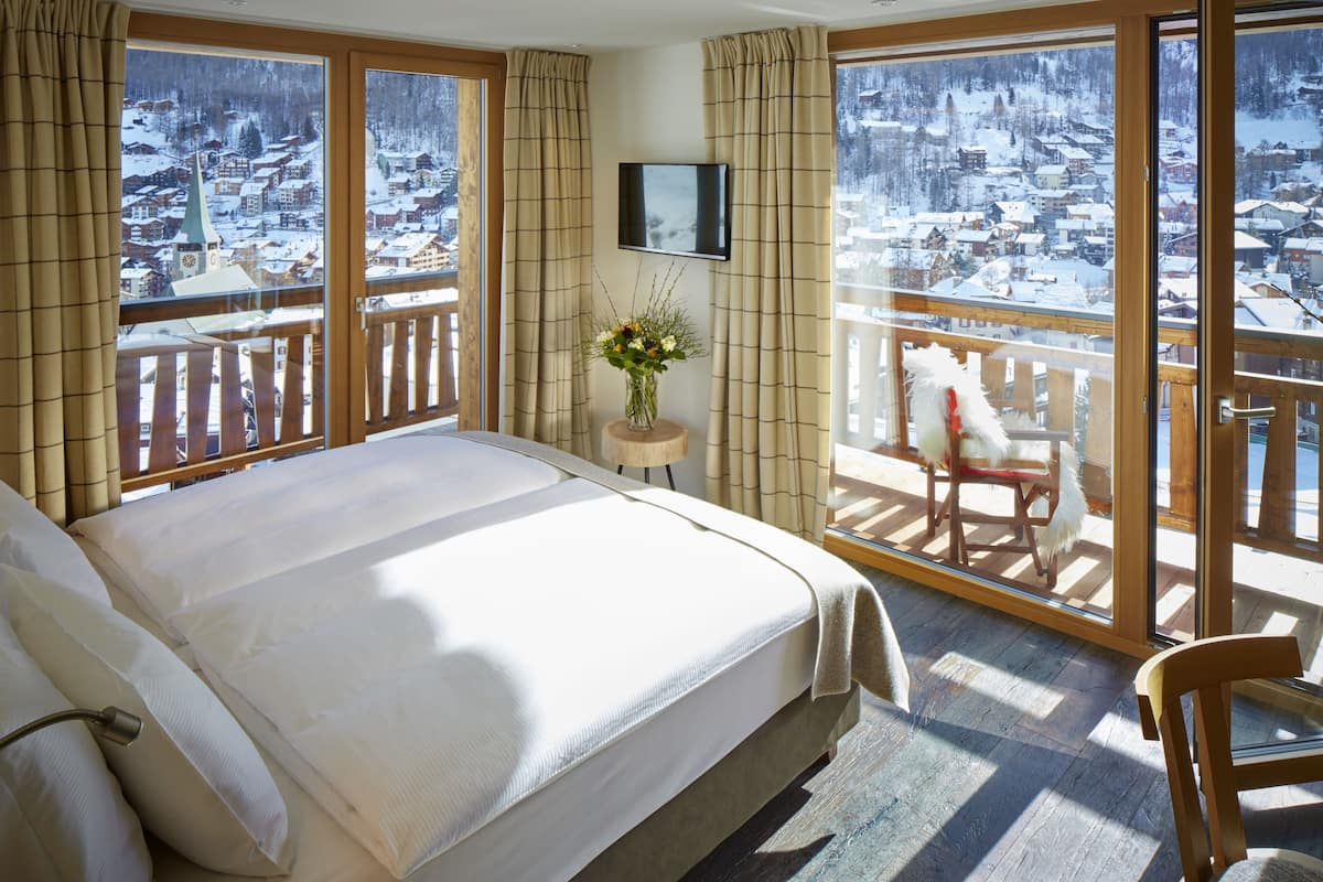 Habitación de hotel con una cama grande, una silla de madera, una mesita con un florero y un televisor en la pared.