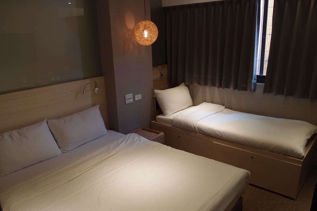 A hotel room with two beds, a bedside table, a lamp, and a window with curtains.