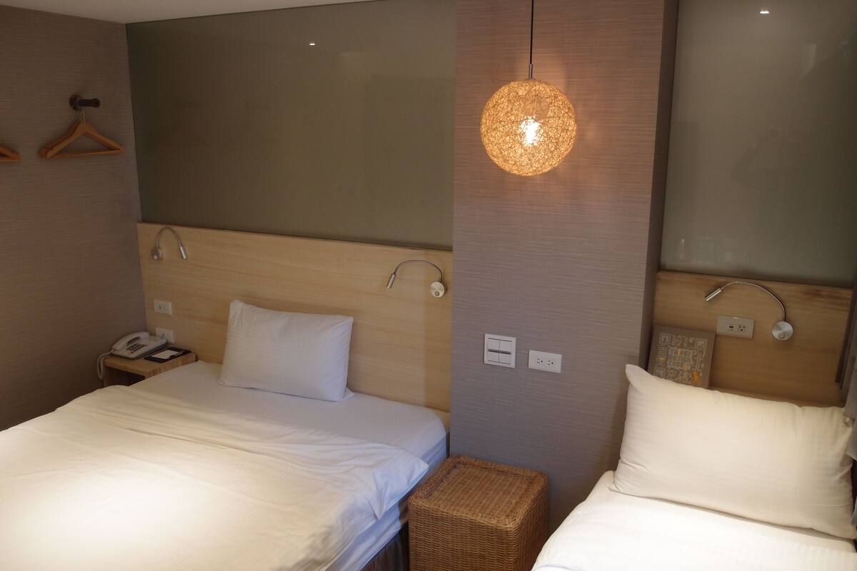 A hotel room with two beds, a wooden headboard, a hanging light fixture, a bedside phone, and a small wicker stool.