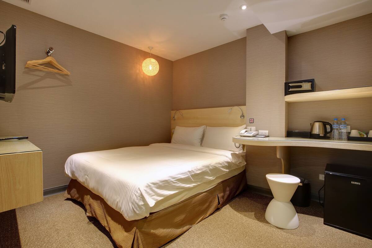 A hotel room with a bed, bedside table, desk, chair, and a small refrigerator.