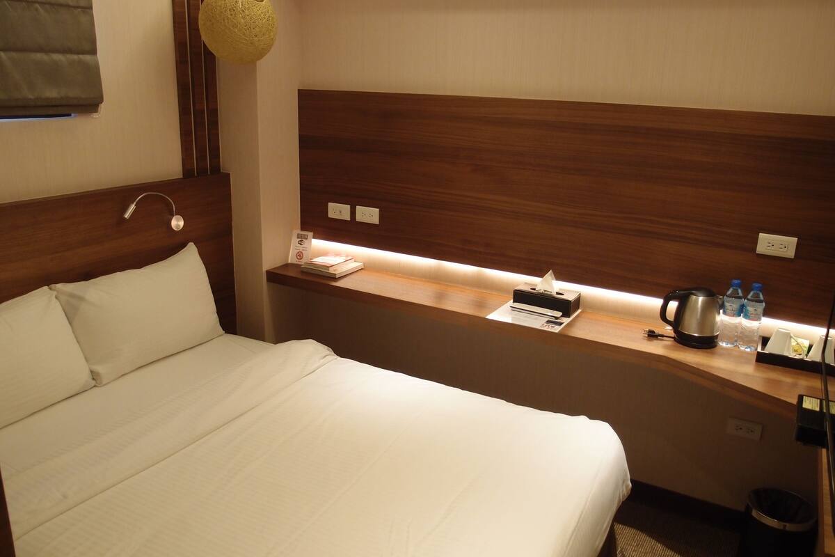 A hotel room with a large bed, a wooden headboard, a bedside table with a lamp, a small shelf with a tissue box, a kettle, and bottled water.