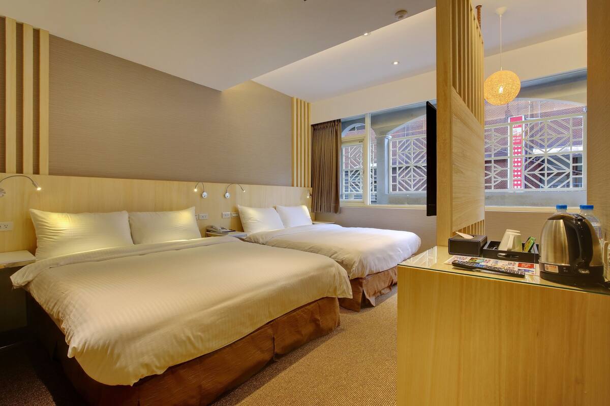 A hotel room with two beds, a desk with a kettle, and a window with a view of a building.