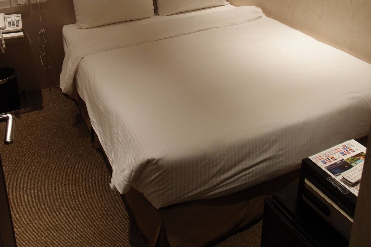 A hotel room with a bed, a nightstand, a lamp, and a small table with a magazine.