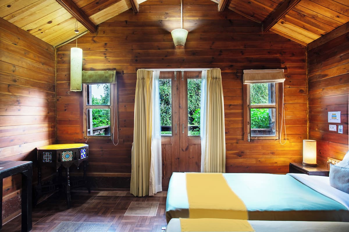 Une pièce de cabane en bois comprenant un lit, deux fenêtres avec des rideaux, une table et un luminaire suspendu.