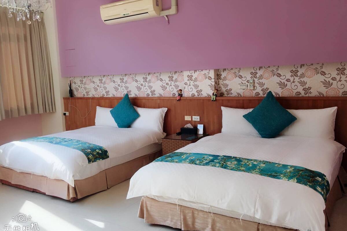 A hotel room with two beds, each with white bedding and teal pillows, a wooden headboard, and a floral wallpaper design.