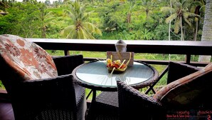 A balcony with wicker furniture and a glass table, overlooking a lush garden with palm trees. - Capung Sakti Villas – By Fair Future Foundation (Ubud)