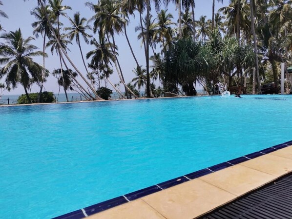 A swimming pool with clear blue water, surrounded by palm trees, and a tiled deck in the foreground. - Helios Beach Resort (Rekawa)