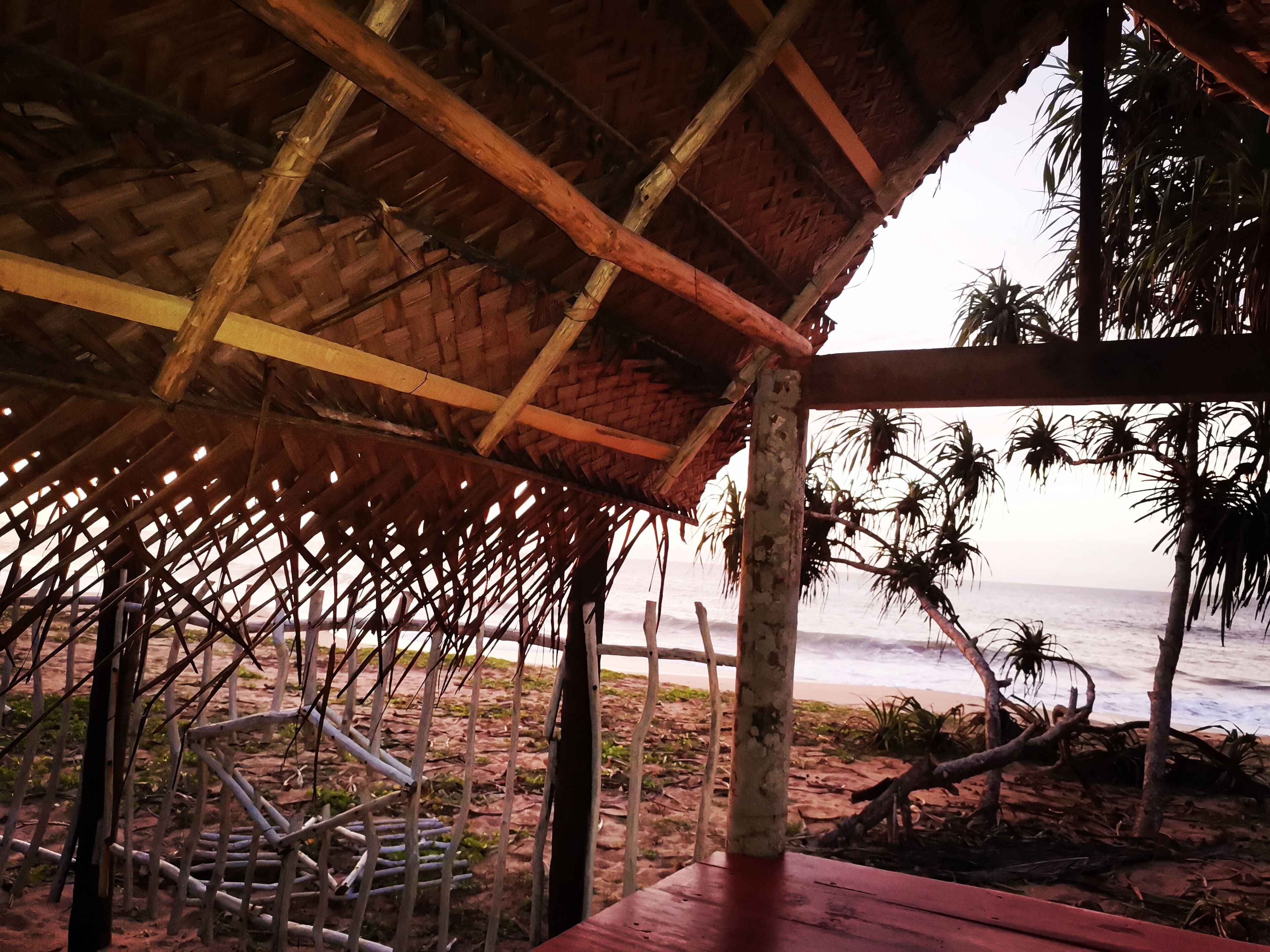 Una cabaña frente a la playa, con techo de paja, vigas de madera y vista al océano.