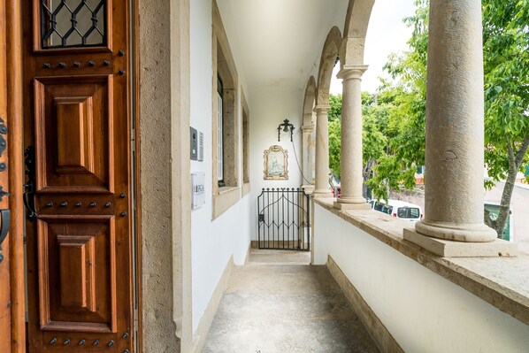 Un couloir avec une porte en bois, des colonnes et un portail donnant sur un espace extérieur où sont garées des voitures et où l'on trouve de la verdure.