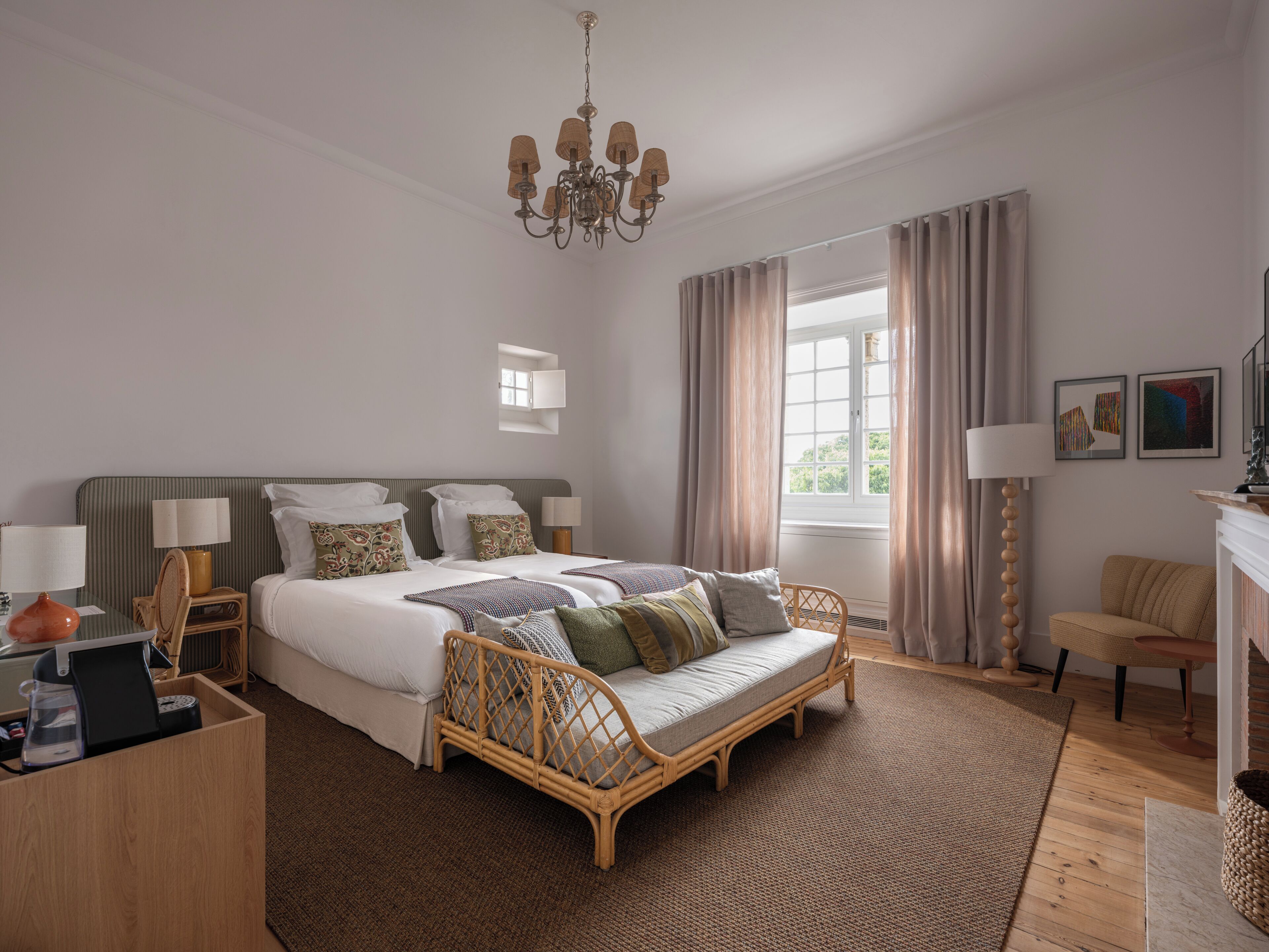 A bedroom with a bed, bedside tables, a bedside lamp, a wicker sofa, a chandelier, a window with curtains, and artwork on the wall.