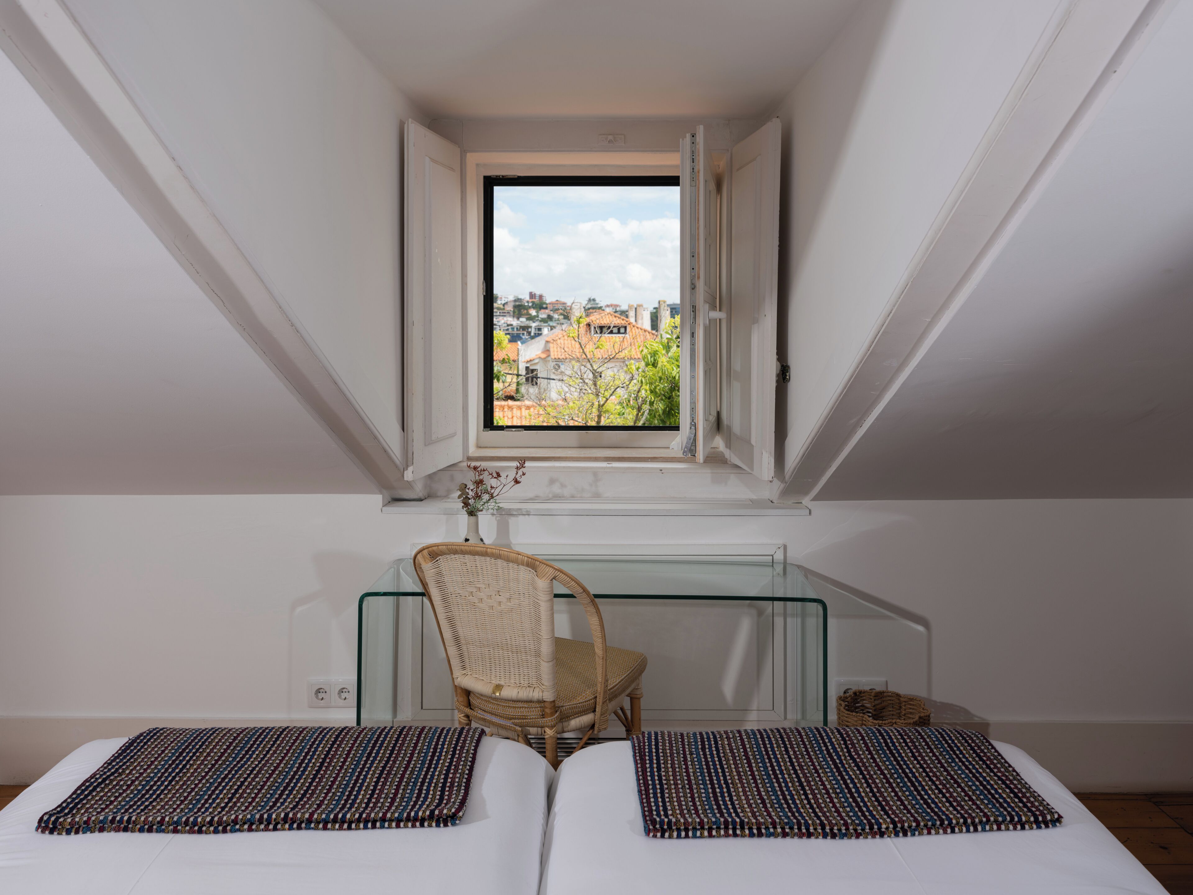 A bedroom with a glass desk, a wicker chair, a bed with two pillows, and a view of buildings through a window.