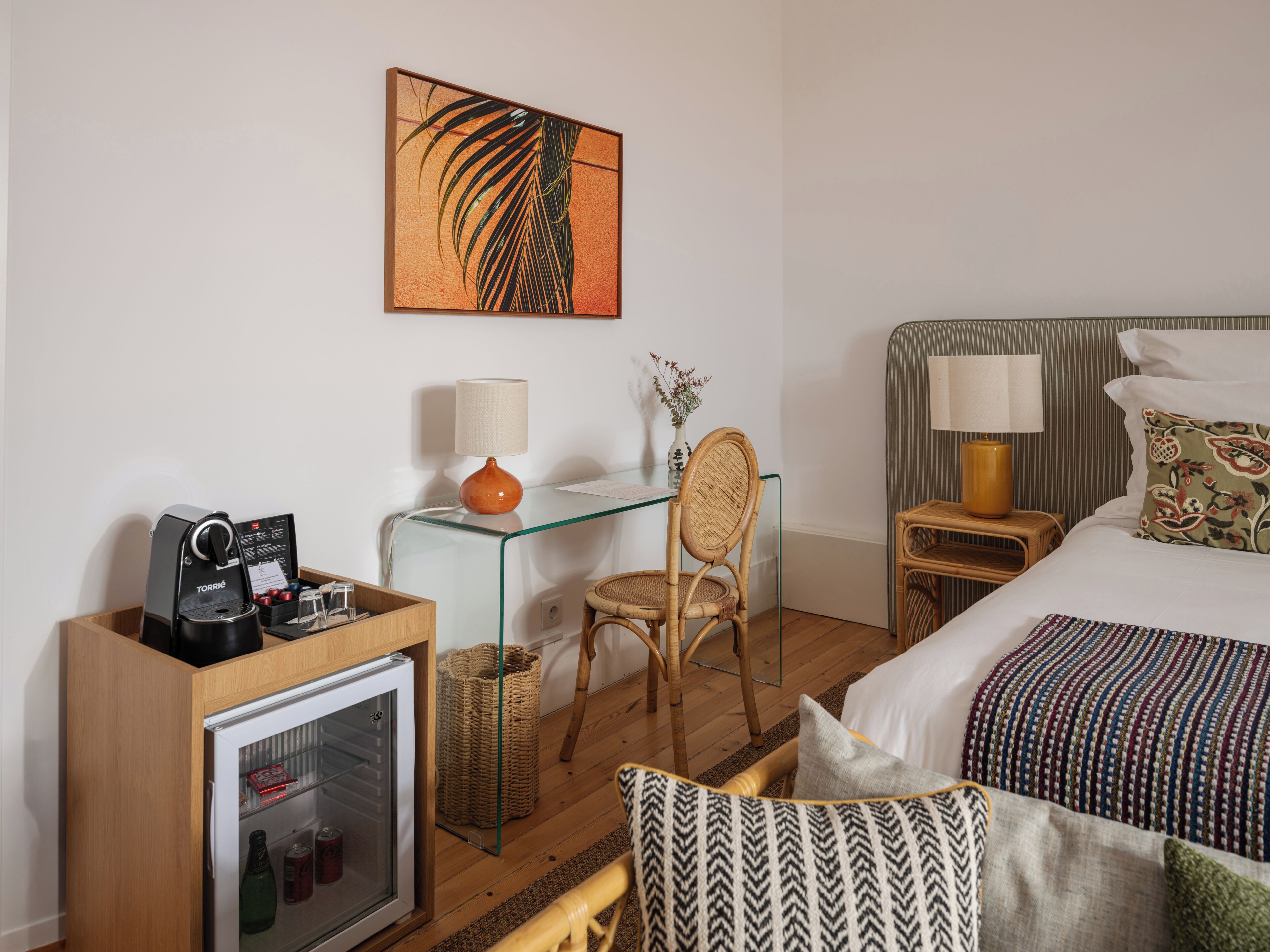 Habitación de hotel con una cama, un escritorio con cafetera, una silla, una mesita con un florero y una obra de arte en la pared.