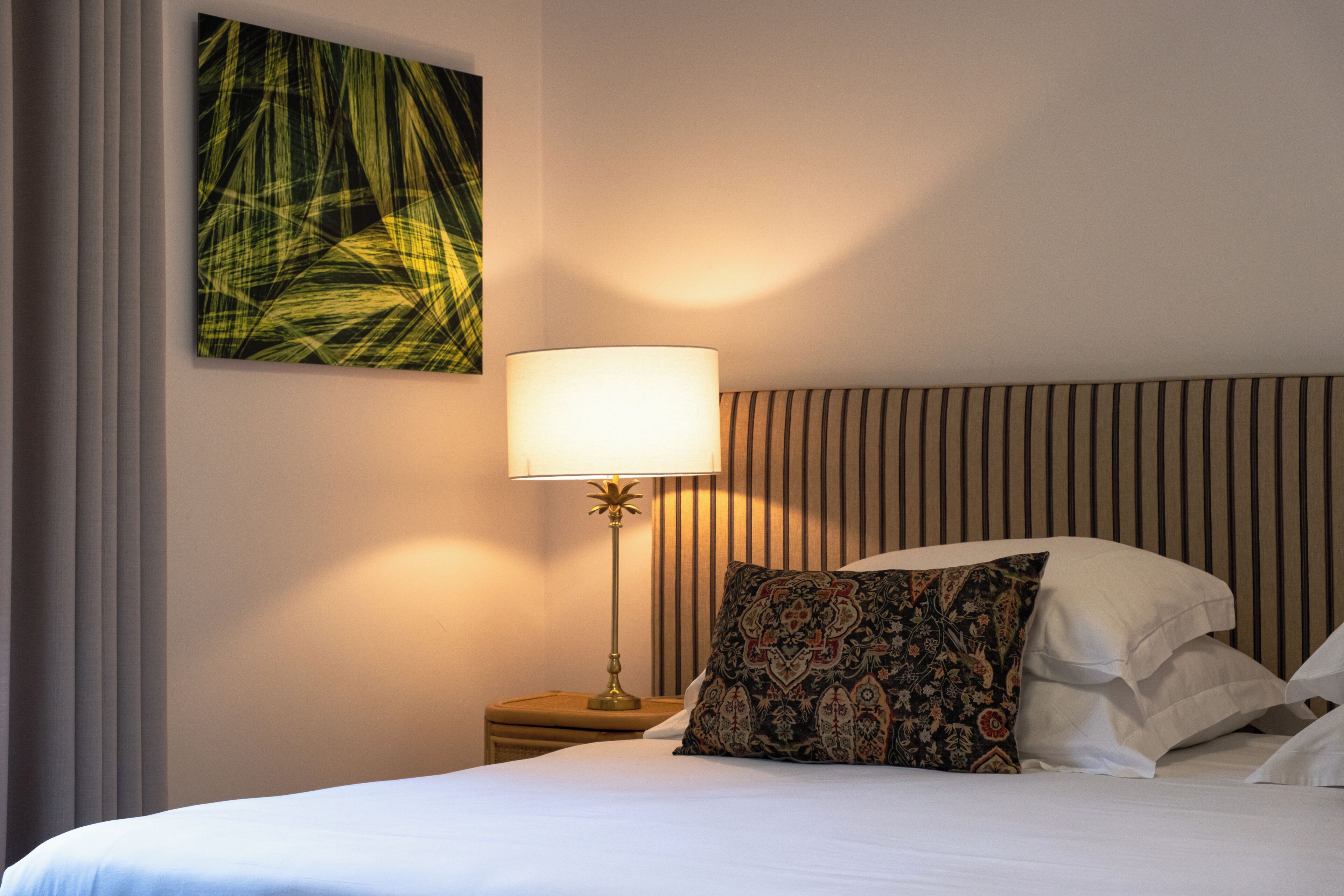 A neatly made bed with white linens, a patterned pillow, a bedside lamp, and a wall-mounted artwork.