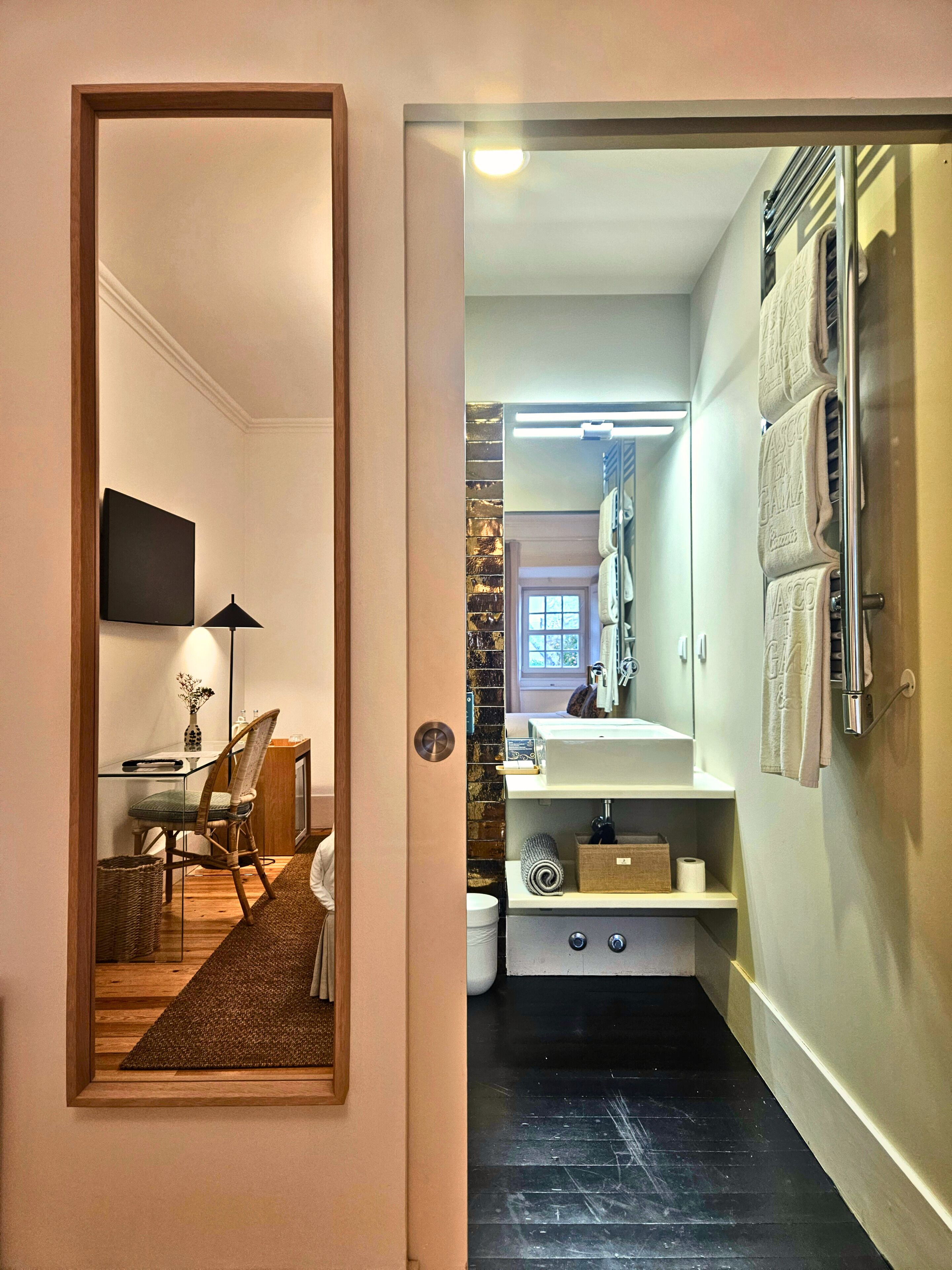 A hallway with a mirror reflecting a dining area, a bathroom with a sink and towel rack, and a door leading to another room.