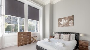 A bedroom with a large bed, a wooden dresser, a window with blinds, and a wall-mounted artwork. - Destiny Scotland - Q-Residence (Edinburgh)