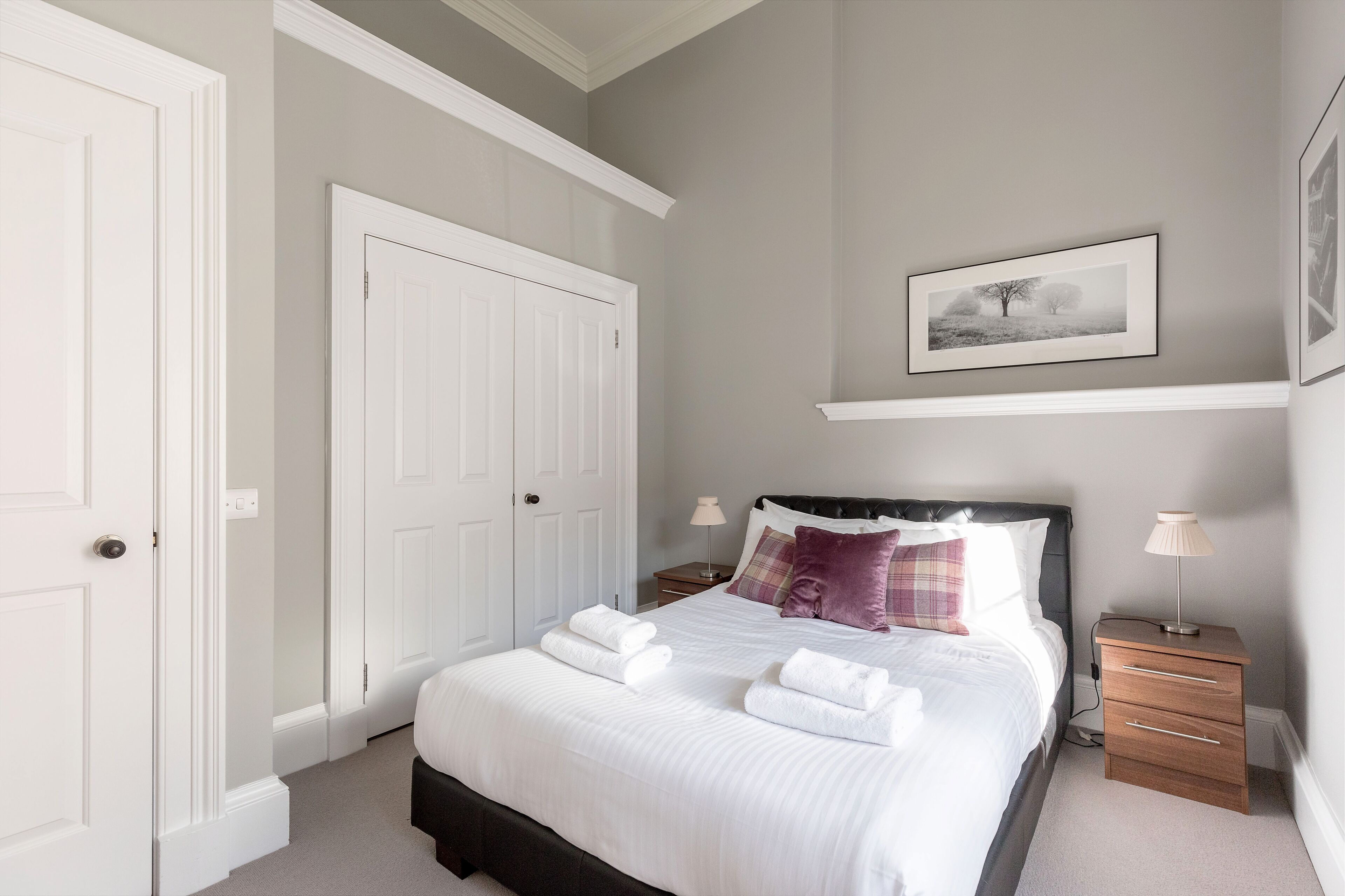A neatly made bed with white linens and two towels, a bedside table with a lamp, and a framed picture on the wall.