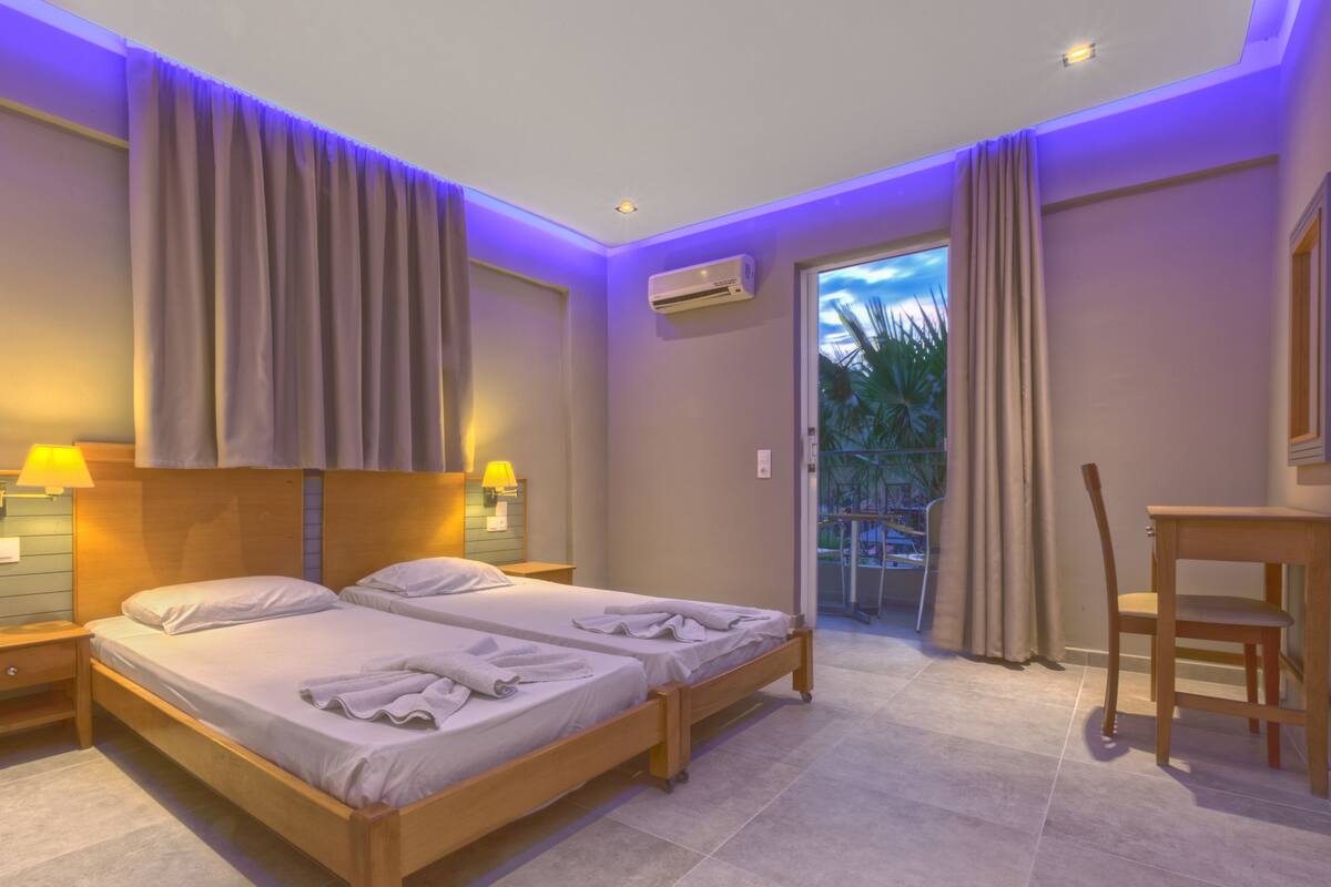A hotel room with a double bed, a desk, a chair, an air conditioning unit, and a view of the outdoors.