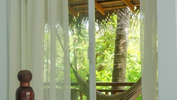 A room with a bed, a wooden sculpture, and a view of a hammock and greenery outside. - Unawatuna Apartments (Unawatuna)