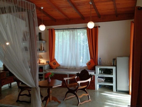A cozy room with a wooden ceiling, a dining table with chairs, a bench with cushions, a small white cabinet, and a window with curtains.