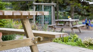 Une table de pique-nique en bois, située dans un jardin, avec une clôture verte et de la verdure en arrière-plan.