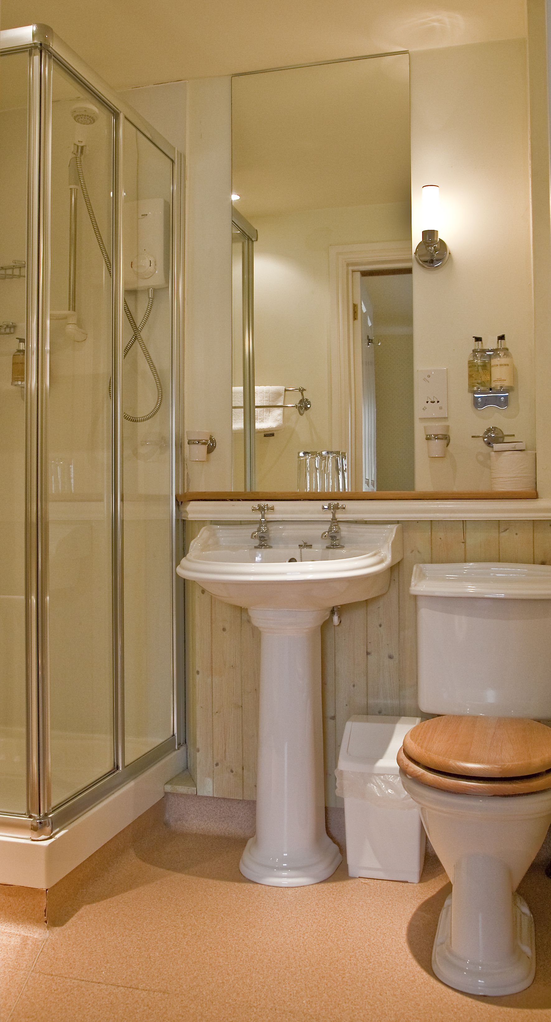 Un baño con ducha de vidrio, lavamanos pedestal, inodoro y un espejo grande.