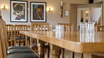 Une longue table à manger en bois, entourée de chaises, avec des œuvres d’art encadrées accrochées au mur et une bouteille de vin.