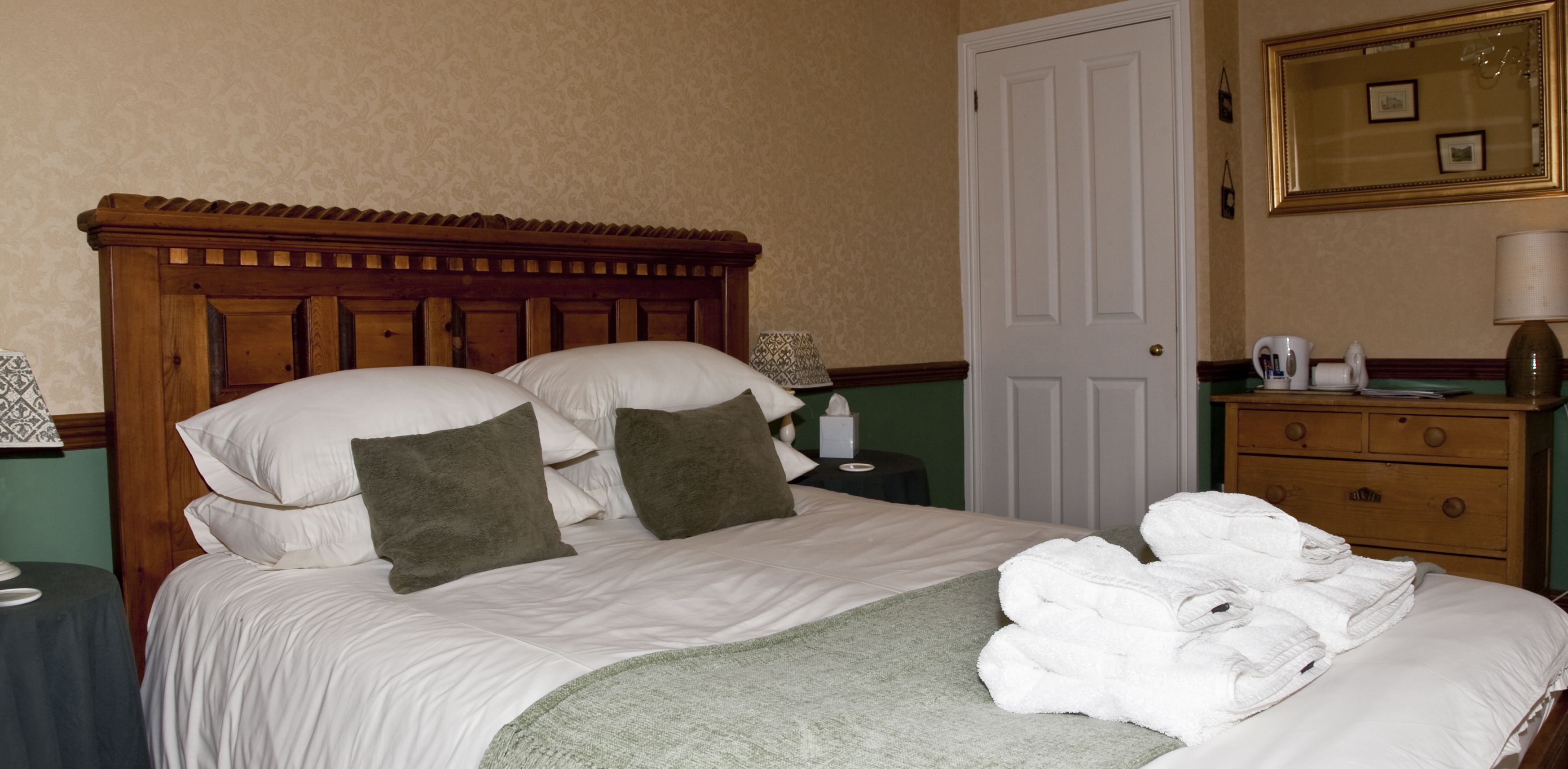 Una cama bien hecha con sábanas blancas, almohadas marrones y una manta verde, junto con una pila de toallas blancas a un lado.