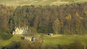 Une grande maison historique, dotée de nombreuses pièces et d’une pelouse bien entretenue, entourée d’une zone boisée et de collines.